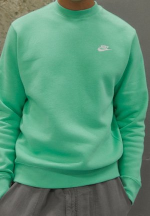 Personne portant un sweat-shirt Nike bleu sarcelle avec le logo sur la poitrine et un pantalon gris, mains dans les poches, sur un fond neutre.