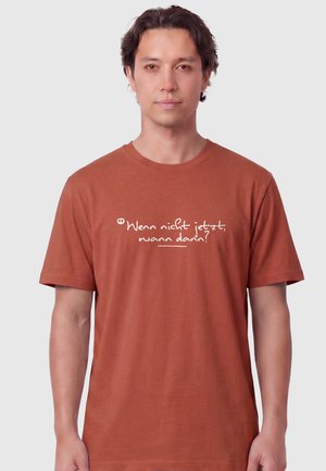 Man with short dark hair wearing a rust-colored t-shirt with the German text "Wenn nicht jetzt, wann dann?" in white script.