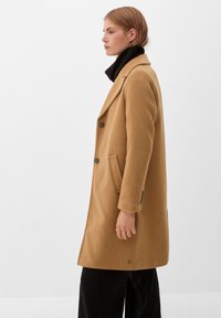 Manteau en laine de chameau avec un col cranté, un design double boutonnage, des poches latérales et des détails en bouton sur les manches et l'ourlet.