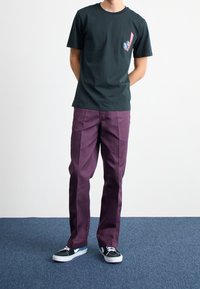 Svart bomullst-shirt med grafiskt tryck, kombinerad med löst sittande, lila byxor och svarta sneakers med vita detaljer.