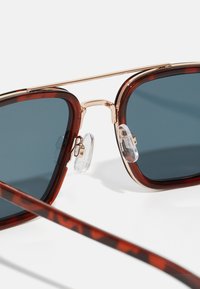 Occhiali da sole tartarugati marroni con lenti sfumate e dettagli in metallo dorato. La montatura presenta naselli trasparenti e un design elegante.
