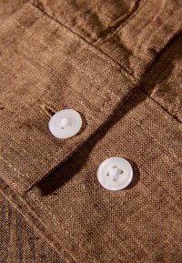 Tessuto marrone con trama e due bottoni bianchi con quattro fori ciascuno, evidenziando cuciture dettagliate lungo i bordi.