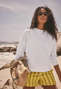 Weißer Sweatshirt mit hohem Kragen, kombiniert mit gelb gestreiften Shorts. Hält eine gemusterte Tasche und steht am Strand.