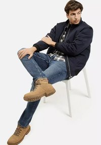 Mężczyzna siedzi na białym krześle, ubrany w granatową kurtkę, koszulę w kratę, niebieskie dżinsy i jasnobrązowe buty na sznurówki, patrząc w bok.