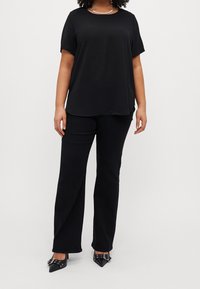 Donna che indossa una maglietta nera a maniche corte, pantaloni neri a gamba dritta e décolleté neri appuntiti con dettagli a fibbia.