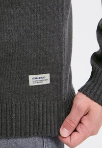 Grå strikket sweater med ribbet kant og manchetter. Har et mærke med "BLEND" tekst, synlige sting og en blød tekstur.