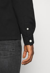 Bras droit en chemise noire à manches longues avec boutons blancs, reposant à côté d'une poche de jean gris foncé.