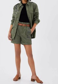 Frau trägt olivgrüne Shorts und passende Jacke mit Knöpfen und Blumenstickerei, schwarzes Oberteil, brauner Gürtel und hellbraune, offene Blockabsatz-Mules.