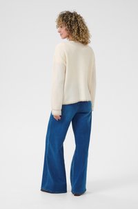 Pulover tricotat de culoare bej deschis, cu mâneci lungi, guler rotund și manșete cu nervuri. Asortat cu blugi din denim albastru, cu picioare largi.