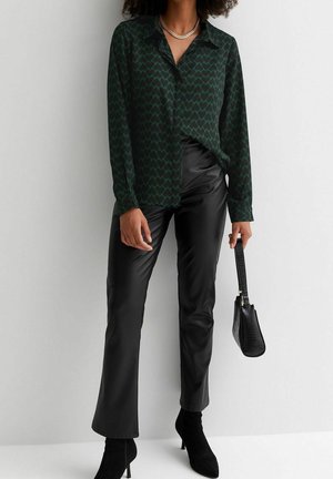 Woman wearing dark green heart-patterned blouse, black leather pants, black heeled boots, holding black handbag against white wall.
