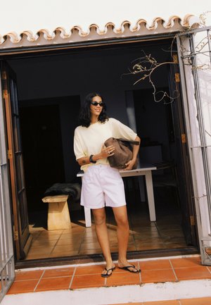 Femme portant des lunettes de soleil, une chemise crème, un short blanc et des tongs, debout dans une porte, tenant un sac marron, souriant à l'extérieur.
