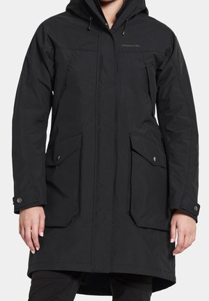 Manteau d'hiver noir à capuche avec fermeture éclair frontale, deux grandes poches à rabat et poignets boutonnés porté par une personne avec les mains détendues sur les côtés.