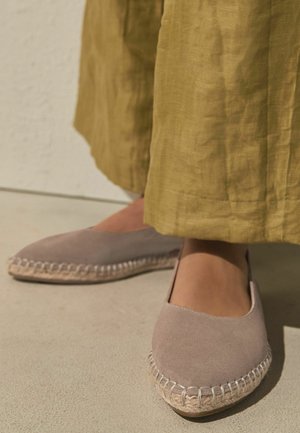Feet wearing beige espadrille flats with stitched jute soles, partially covered by loose mustard-colored linen pants on a light floor.