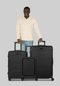 Tre valigie rigide nere di diverse dimensioni, dotate di manici telescopici, texture lisce e sottili linee orizzontali.