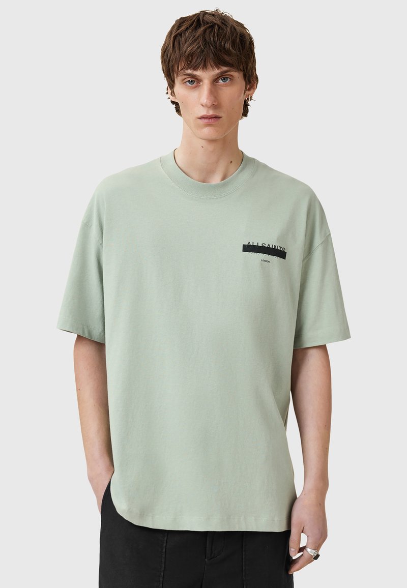 Camiseta de algodón verde claro con cuello redondo, mangas cortas y un logotipo negro impreso en el lado izquierdo del pecho. Textura suave y corte holgado.