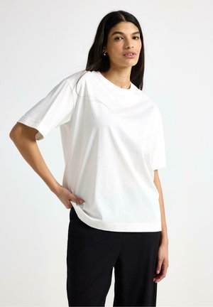 Kvinde med langt mørkt hår iført en enkel hvid t-shirt og sorte bukser, stående med den ene hånd på hoften mod en hvid baggrund.