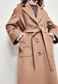 Abrigo de lana beige con cintura ceñida, solapas con muesca, tres botones grandes y dos bolsillos frontales, que presenta una textura suave y un corte holgado.