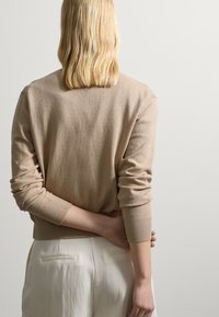 Beige pletený kardigan s kulatým výstřihem a žebrovanými manžetami, v kombinaci se světlými kalhotami, zobrazený z zadní strany.