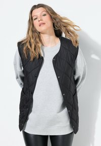 Schwarze gesteppte Weste mit Druckknöpfen, die über einem grauen Sweatshirt getragen wird. Sie hat eine entspannte Passform und eine glatte Textur. Betont den Lagenlook.