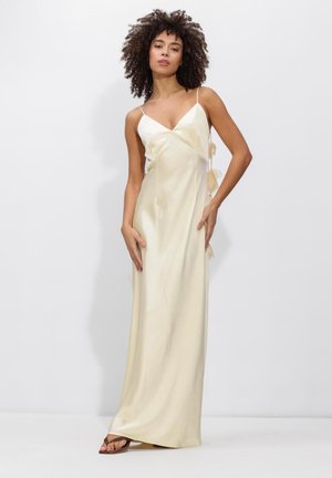 Vrouw met krullend haar, gekleed in een lange, roomkleurige slipdress met spaghetti bandjes, staand tegen een effen witte achtergrond.