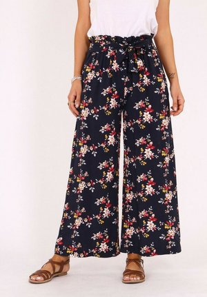 Pantalon large bleu marine avec imprimés floraux blancs, rouges et jaunes, taille paperbag, ceinture nouée, porté avec un haut blanc et des sandales marron.