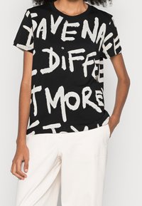 Czarny t-shirt z wyraźnym białym napisem o różnych rozmiarach, krótkimi rękawami i okrągłym dekoltem, zestawiony z jasnymi spodniami.