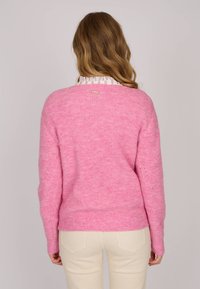 Pull en tricot rose avec une texture douce, coupe ample, manches longues et un détail de marque subtil dans le dos. Porté par-dessus une chemise à col.