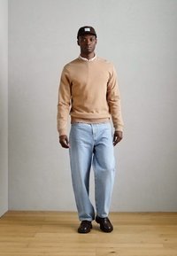 Sweatshirt beige chiaro con polsini a coste, jeans in denim blu e scarpe marrone scuro. Il modello indossa un cappellino marrone con una toppa bianca con il logo.