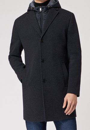 Homme portant un manteau en laine gris foncé boutonné par-dessus un sweat à capuche noir zippé, associé à un jean bleu, sur un fond clair uni.