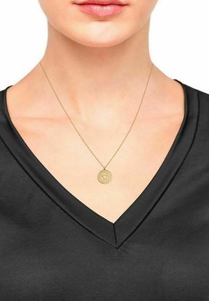 Collana dorata con un pendente rotondo con dettagli intricati, indossata su una maglietta nera a scollo a V. Texture del tessuto liscia visibile.