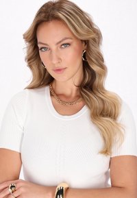 Witte geribbelde shirt met korte mouwen heeft een ronde hals en kabelbrei-details. Een gouden ketting en een opvallende metalen armband worden gedragen.