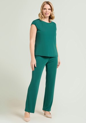 Donna in piedi, indossa un top verde senza maniche e pantaloni abbinati, con scarpe beige con tacco alto, sfondo neutro.