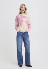Pull en mélange de laine couleur crème avec des motifs de feuilles roses, associé à un jean bleu à jambes larges. Le pull présente un col rond et des poignets côtelés.
