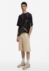 Crna košulja kratkih rukava s šarenim apstraktnim linijama; bež kratke hlače; bijele i narančaste tenisice; jednobojna pozadina.