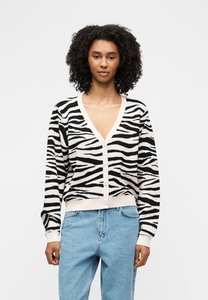 Kobieta o kręconych włosach, ubrana w czarno-biały kardigan w paski zebry zapinany na guziki oraz jasnoniebieskie dżinsy, stojąca na tle jednolitego białego tła.
