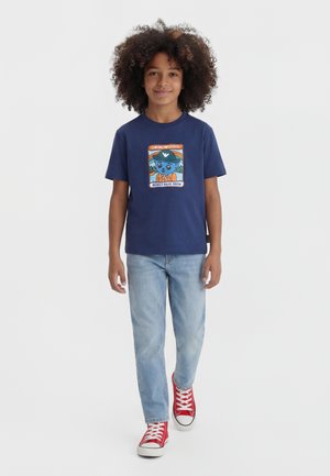 Barn med krøllet hår har en mørkeblå t-shirt med grafik, lyseblå jeans og røde sneakers på, og går fremad mod en hvid baggrund.