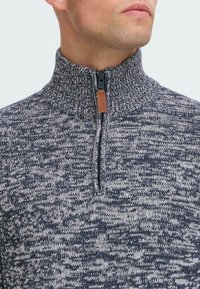 Hombre vistiendo un suéter de punto jaspeado en gris y azul marino con un cuello alto acanalado y una cremallera frontal con un tirador marrón.