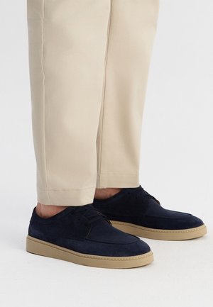 Granatowe zamszowe buty z okrągłym noskiem, sznurowane, z beżową gumową podeszwą, zestawione z beżowymi spodniami, prezentujące casualowy styl.