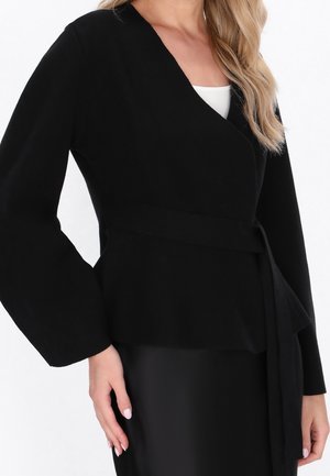 Femme portant une veste noire style cache-cœur avec une ceinture nouée à la taille et un haut blanc en dessous, montrant le haut du torse et les bras.
