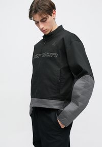 Casaco bomber preto com uma textura suave, apresentando uma fecho de correr à frente, manga cinza acolchoada e logótipo bordado no peito.