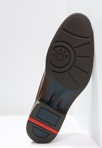 Semelle de chaussure en cuir marron avec un motif texturé. Présente un logo circulaire en relief et des accents bleus avec une bande rouge sur le talon pour l'adhérence.