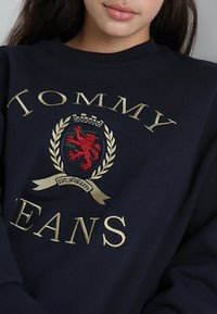Marinblå sweatshirt med guldstickning av "TOMMY" och "JEANS," med en röd lejonkrona och lagerkransdetaljer. Slät yta, rundad hals.