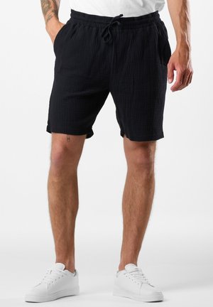 Shorts en coton noirs avec une taille élastique et un cordon de serrage, dotés d'un tissu texturé et de poches latérales, portés avec des baskets blanches.