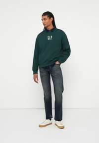 Felpa verde con logo "GAP" sul petto, abbinata a jeans scuri e scarpe bianche con suola beige, in piedi davanti a uno sfondo neutro.