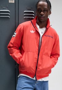 Röd, lättviktsjacka med dragkedja och elastisk nederkant, med vit Adidas-logotyp på bröstet och en fotbollsdesign på ärmen. Bärs över en vit skjorta.