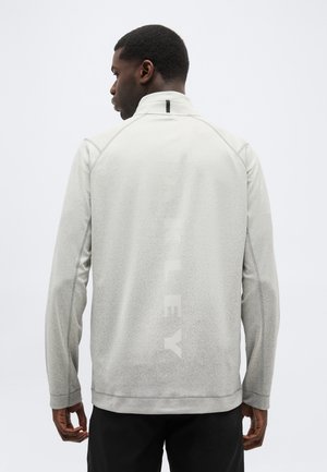 Hombre de espaldas vistiendo una chaqueta gris claro con la palabra "HURLEY" débilmente visible en vertical en la parte trasera, contra un fondo liso.