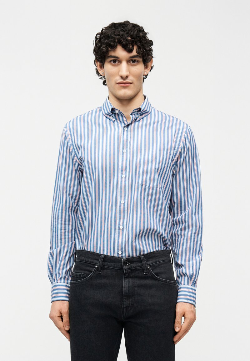 Νεαρός άνδρας με κοντά σγουρά μαλλιά φοράει πουκάμισο με κάθετες ρίγες σε μπλε, λευκό και ροζ χρώμα και μαύρο τζιν, στέκεται μπροστά σε λευκό φόντο.