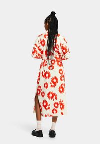 Vestido maxi floral con fondo crema, que presenta grandes flores rojas, mangas largas y aberturas laterales. Combinado con zapatos negros y calcetines a rayas.