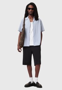 Camicia azzurra chiaro a maniche corte con bottoni sopra una canotta bianca, abbinata a pantaloni corti neri e scarpe nere lucide. Borsa marrone a motivo e occhiali da sole.
