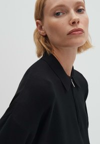 Indumento nero lavorato a maglia con colletto, dotato di chiusura con zip e texture a coste. Il modello ha capelli corti e uno sfondo neutro.